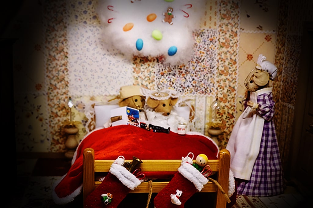 Two baby mice snuggled into bed with a cloud of candy overhead and stockings attached to the footboard.