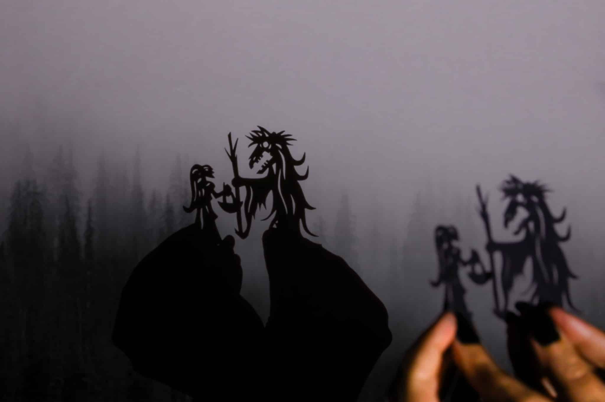 Tania Yager holding two small cut-paper shadow puppets which cast shadows onto a misty forest scene.