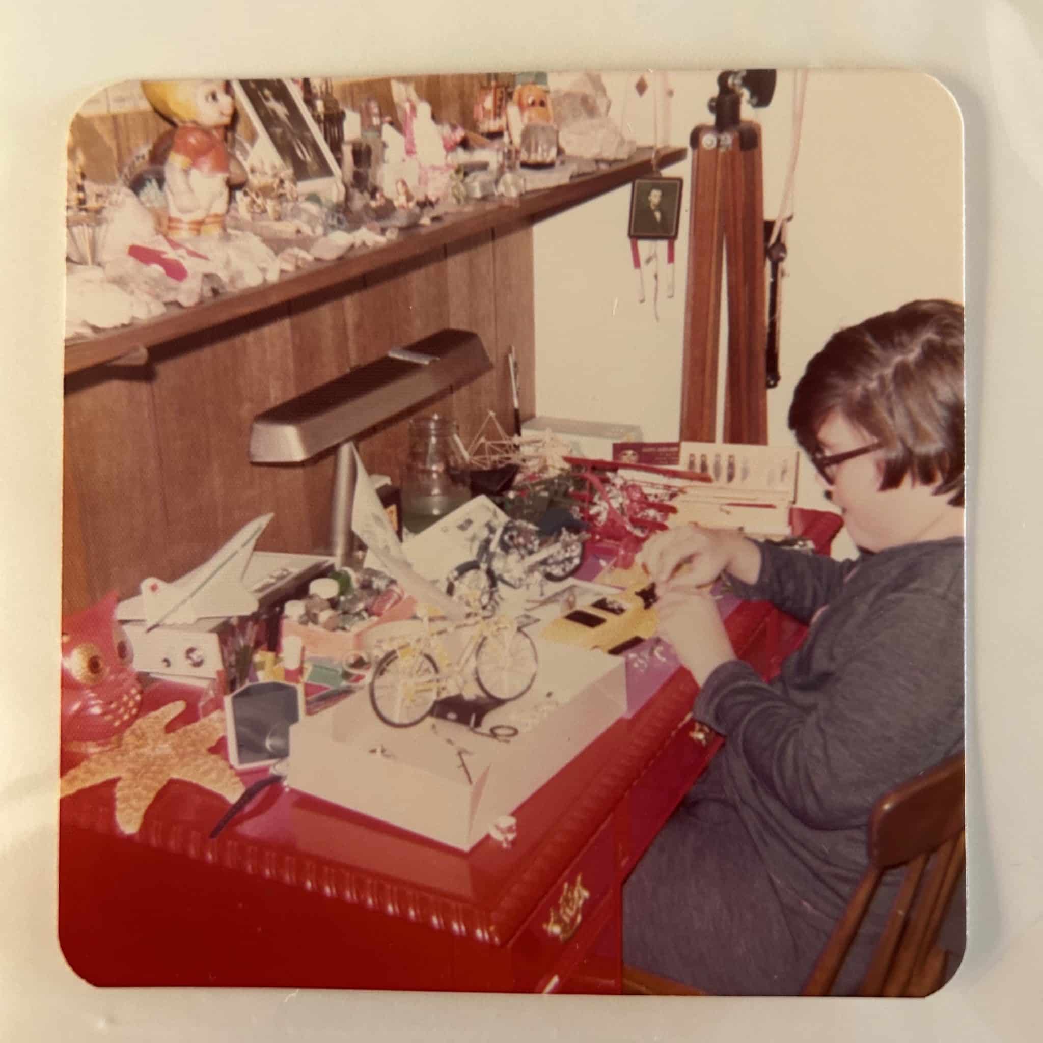 Stephen Farley building scale models in his bedroom, age 11. On his desk are scale models of a bicycle, motorcycle, old car, Concorde jet, and a Fokker triplane, along with various paints and X-Acto knives.