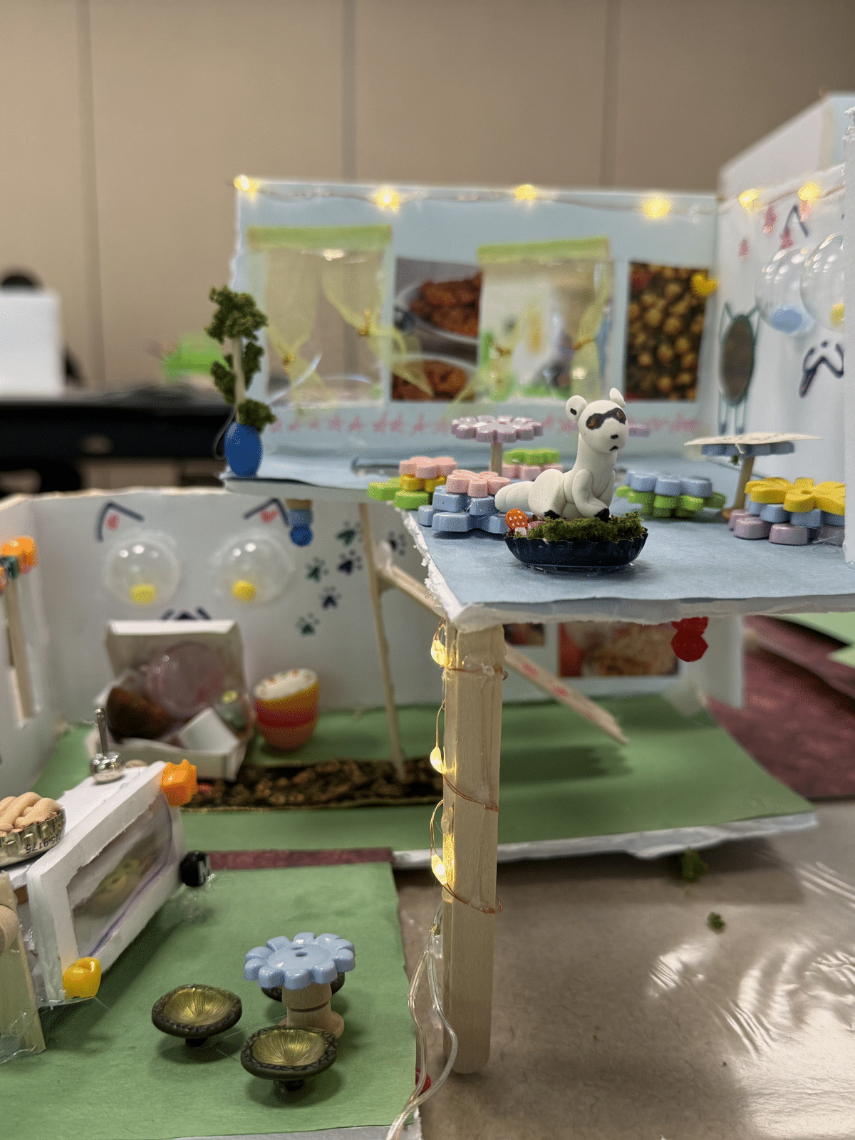 Miniature diorama featuring a white dog figurine on a blue platform amid pastel furniture and string lights in a cozy display.